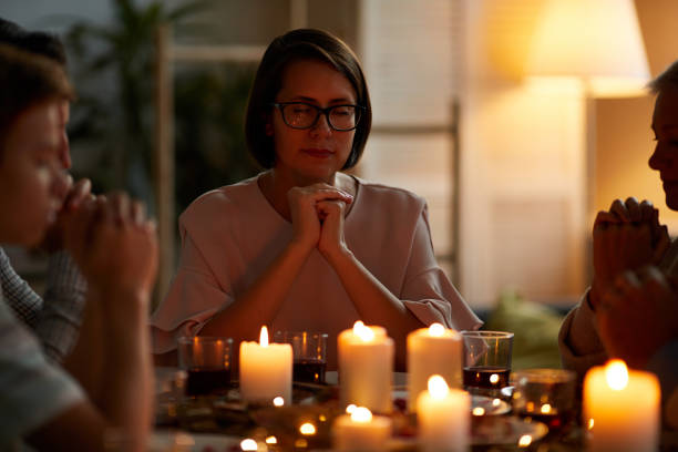 Young woman in eyeglasses sitting at the table with candles together with her family and they praying before dinner