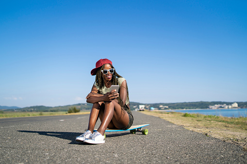 Woman sitting on skatebosrd and using smart phone, on road.