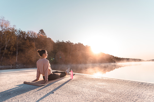 A young woman is exercising yoga by a lake early in the morning on a beautiful autumn day. She is leaning back, enjoying the sun.