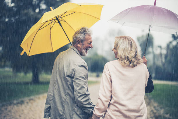 back view of happy senior couple with umbrellas taking a walk on rainy day. - umbrella senior adult couple autumn imagens e fotografias de stock