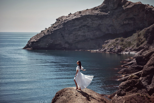 A beautiful girl, bride, in a white dress stands on a cliff against the background of the sea and mountains, her dress develops from the wind. With makeup and hairstyle. Fashion, style, wedding. Space.