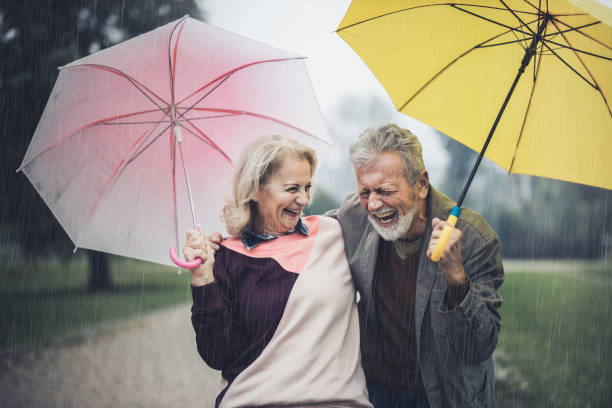 cheerful senior couple with umbrellas on rainy day in nature. - umbrella senior adult couple autumn imagens e fotografias de stock