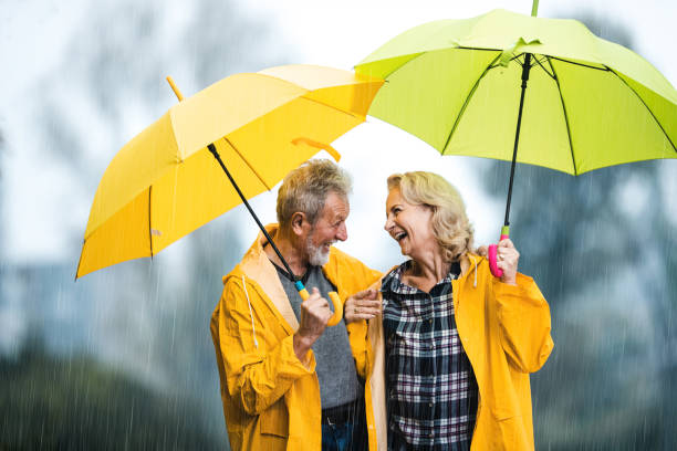 happy senior couple in raincoats talking under umbrellas on a rainy day. - umbrella senior adult couple autumn imagens e fotografias de stock