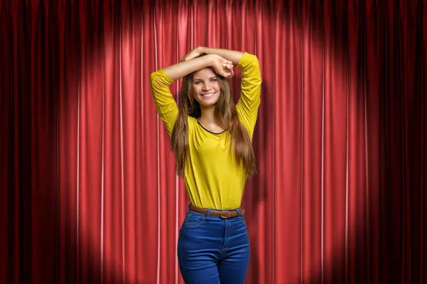 Photo of Front crop view of young smiling girl in yellow jumper and blue jeans, standing in spotlight with arms raised and hands on head, against red stage curtain.