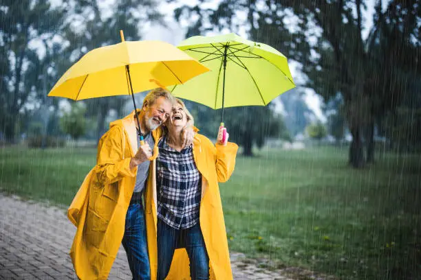 Happy senior couple in raincoats taking a walk under umbrellas on a rainy day at the park.