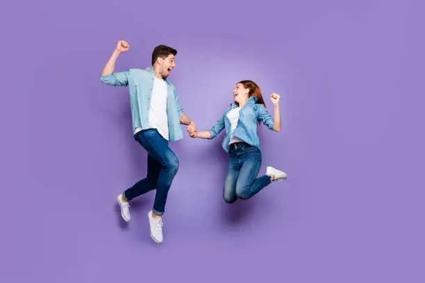 Full size profile side photo of delighted funny brunette hair redhair spouses, jump raise fists scream yeah feel lucky hold hand wear denim jeans shirt sneakers isolated over purple color background