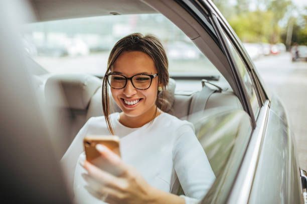 verticale d'une belle femme d'affaires utilisant un téléphone intelligent et souriant tout en s'asseyant sur le siège arrière de la voiture. - passager photos et images de collection