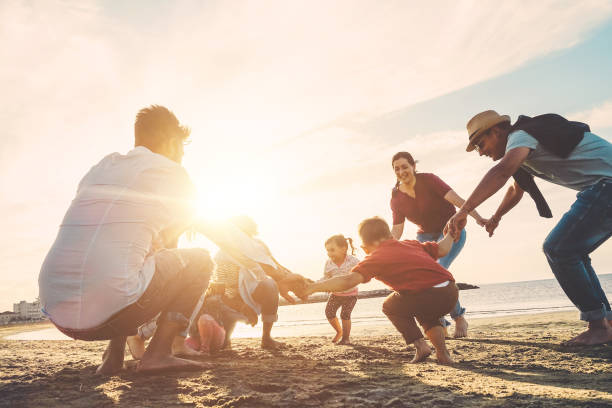 amici di famiglia che si divertono sulla spiaggia al tramonto - padri, madri, bambini e zii che giocano insieme - focus sui corpi - amore, festa e concetto celebrante - soft focus sul viso dell'uomo destro - sunset day back lit autumn foto e immagini stock