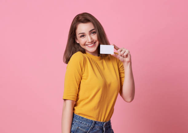 Portrait of a young woman in yellow shirt showing credit card and looking away at copy space isolated over pink background. stock photo