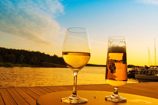 A glass of white wine and beer on the table on the pier at sunset light over ocean sky, with sailboats background.