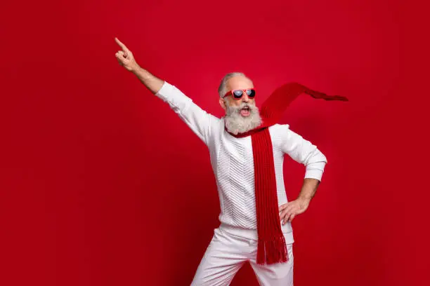 Cool santa character aged man directing finger on advert banner wear, sun specs knitted clothes isolated red background