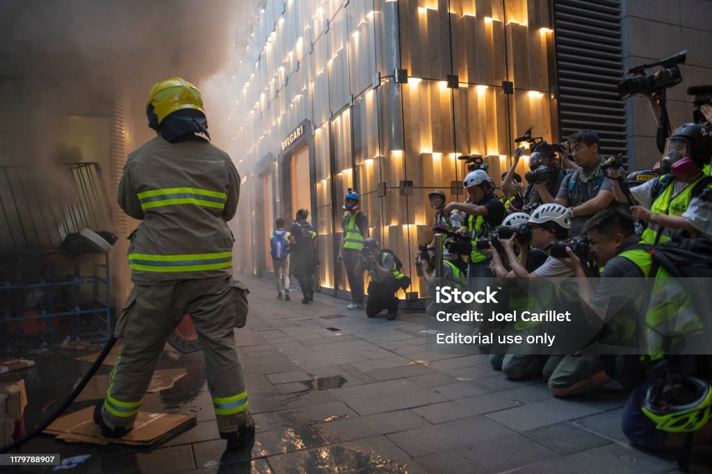 Photojournalists and fireman at work in Hong Kong A fireman extinguishes a blaze at the entrance to Central station in Hong Kong as a large group of journalists, mostly photojournalists, document the scene. (September 8, 2019) Protest Stock Photo