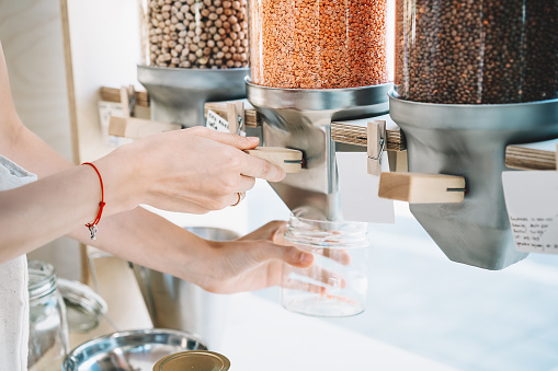 Sustainable shopping at small local businesses. Close-up image of woman pours red lentils in glass jar from dispensers in plastic free grocery store. Girl with cotton bag buying in zero waste shop.