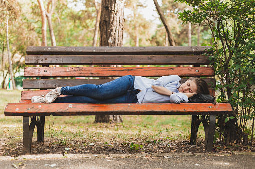 Beautiful young woman sleeping on a bench in the park - Tired female worker wearing jeans and blue shirt taking a nap lying outdoors