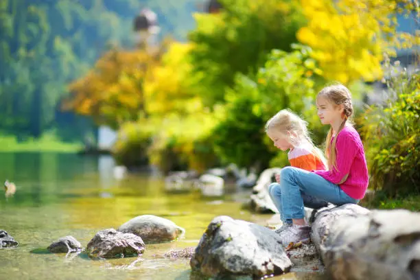 Adorable sisters playing by Konigssee lake in Germany on warm summer day. Cute children having fun feeding ducks and throwing stones into the lake. Summer activities for kids.