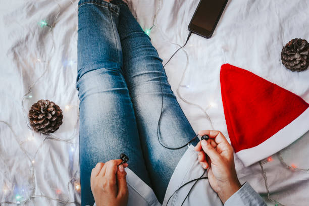 féminin écoutant la musique dans le lit. cônes d'arbre de pin, chapeau de santa claus et smartphone avec des écouteurs près des jambes de femelle. les vacances de noel. le matin de la x-mas. après-midi paresseux. - christmas tree audio photos et images de collection