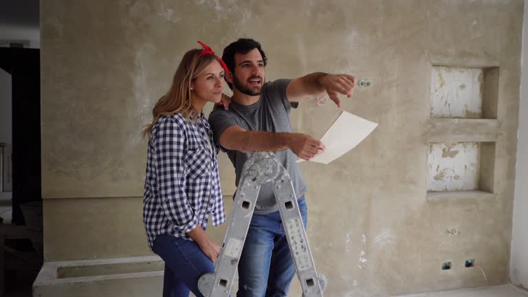 Happy couple doing a home renovation looking at a design on paper while pointing at a wall and talking