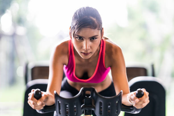 Beautiful Asian woman exercising on stationary cycling machine in indoor fitness gym, determination face. Sport recreational activity, people workout, or healthy lifestyle concept. stock photo