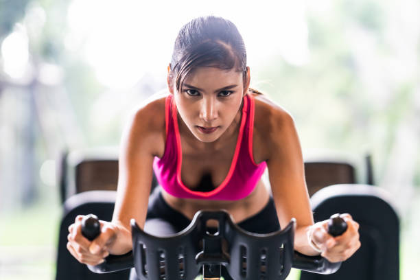 beautiful asian woman exercising on stationary cycling machine in indoor fitness gym, determination face. sport recreational activity, people workout, or healthy lifestyle concept. - bicycle women cycling gym imagens e fotografias de stock