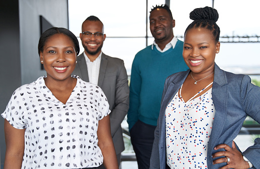 Group of four black professionals wearing fashionable corporate clothing standing confidently together in bright modern office space