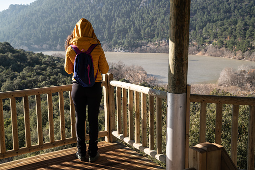 Rear view of adult woman looking at the view from observation point in National Park of Kovada, Isparta, Turkey. Model is wearing a yellow coat and tight pants. She has a small backpack. Shot in day time with a full frame mirrorless camera.