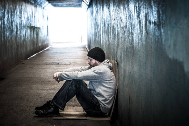 Homeless young man sitting in cold subway tunnel Desaturated color image depicting a profile view of a homeless young male, in his 30s, sitting in a cold and dark subway tunnel. The man looks desperate, sad and forlorn. The tunnel recedes into the distance beyond the man, and we can see the glowing light at the end of the tunnel. Room for copy space. homeless person stock pictures, royalty-free photos & images