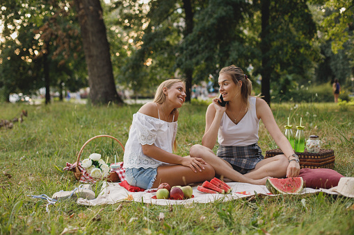 Two young women sitting on  a blanket in the park. They are having healthy snacks and drinks.