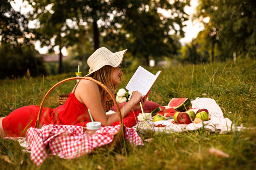 Side view of a beautiful woman wearing red dress and sunhat reading a book while relaxing in the park.
