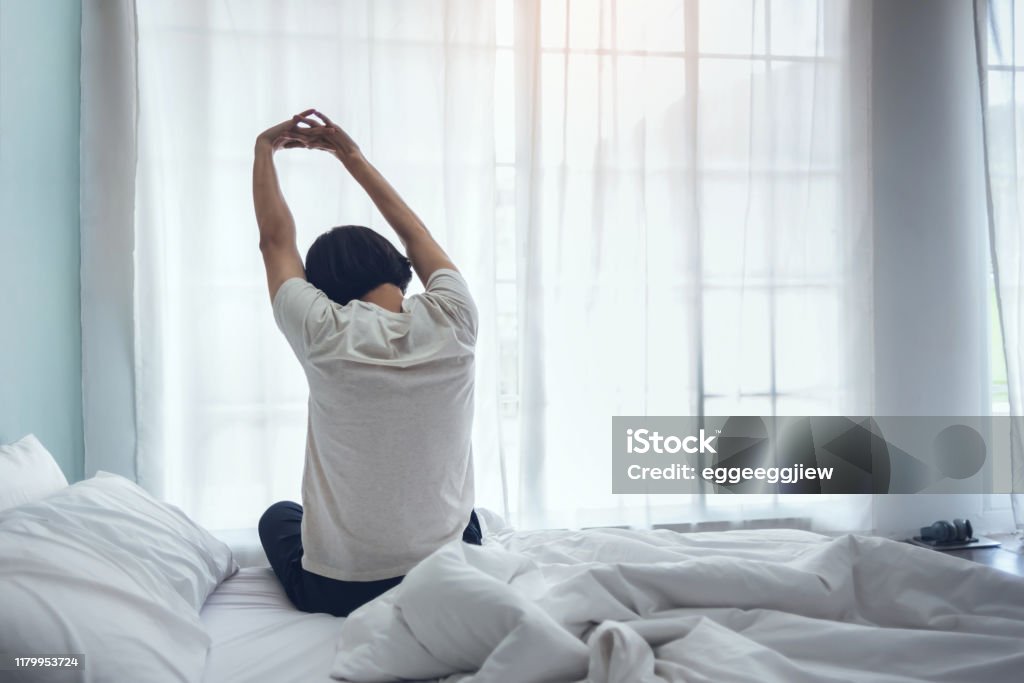 Asian man sitting on the bed and stretching his arms during early morning. Young Asian man sitting on the bed and stretching his arms during early morning at his home with copy space. Holiday, Weekend, Vacation, Good health, Rear view, Wake up. Hands raised in the air. Stretching Stock Photo