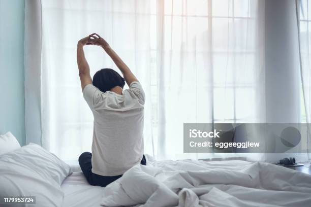 Asian Man Sitting On The Bed And Stretching His Arms During Early Morning Stock Photo - Download Image Now