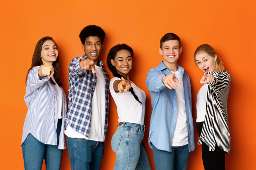 Hey, you! Happy teenagers pointing fingers at camera and smiling over orange background
