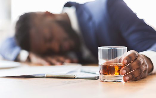 Alcoholism, drinking concept. Drunk african employee sleeping at working place, holding glass with alcohol, selective focus, panorama