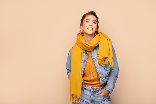 Young beautiful happy girl posing in studio wearing fashionable scarf and jeans jacket. Smiling woman looking at camera. Human emotions,, expression. Beige studio background.