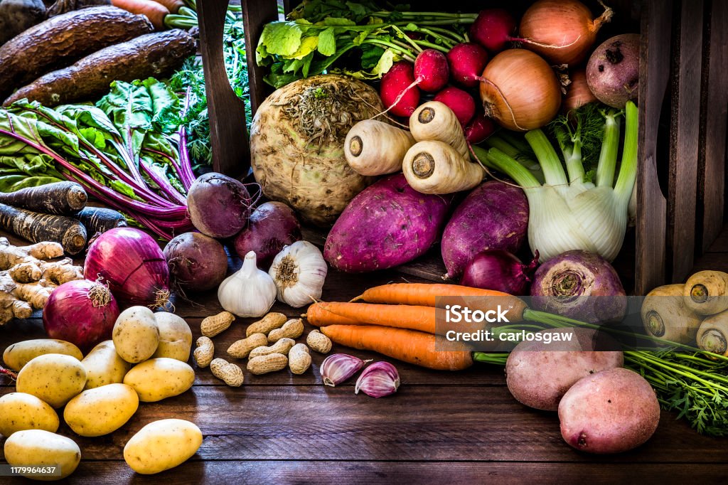 Healthy food: organic roots, legumes and tubers still life. Front view of a large group of multicolored fresh organic roots, legumes and tubers shot on a rustic wooden background. The composition includes potatoes, Spanish onions, ginger, purple carrots, yucca, beetroot, garlic, peanuts, red potatoes, sweet potatoes, golden onions, turnips, parsnips, celeriac, fennels and radish. Some elements are on a rustic wooden crate. Low key DSLR photo taken with Canon EOS 6D Mark II and Canon EF 24-105 mm f/4L Root Vegetable Stock Photo