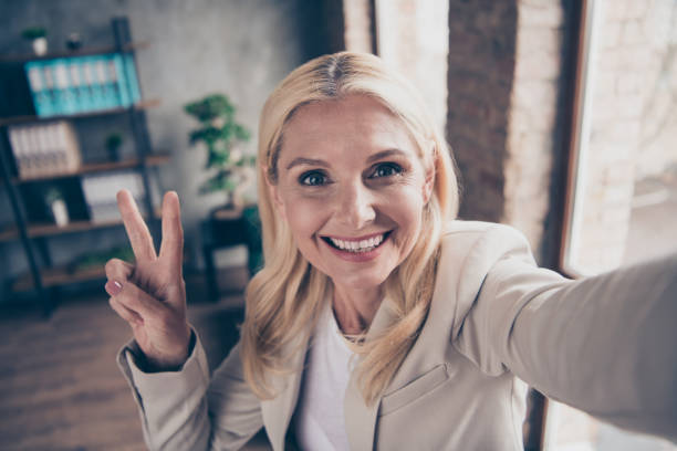 fermez-vous vers le haut de la photo de la main-d'œuvre gaie gaie de femme d'affaires d'âge moyen font le selfie saluent ses clients des investisseurs de partenaires font v-sign dans le bureau d'entreprise de main-d'oeuvre - symbols of peace photos et images de collection