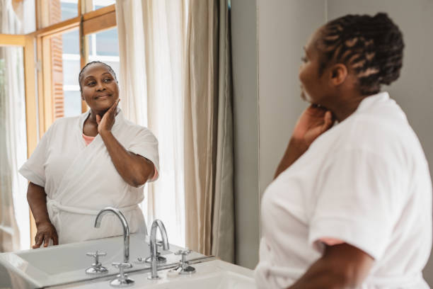 mujer afroamericana mirando su tez en un baño - human face women mirror touching fotografías e imágenes de stock