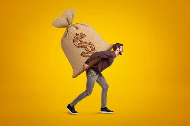 Photo of Side view of young handsome man in casual clothes carrying huge heavy sack with dollar symbol on.