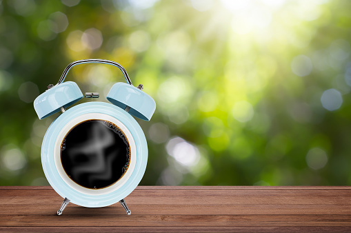 Coffee cup with classic blue alarm clock on wooden table on blurred green nature background