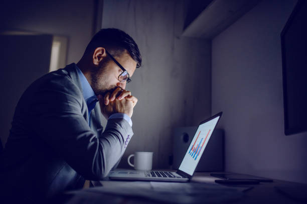 serio bell'uomo d'affari caucasico in abito appoggiato alla scrivania e guardando le classifiche. concetto di lavoro a tarda notte. - sforzo foto e immagini stock