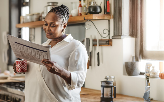 Smiling African American woman drinking a cup of coffee and reading the newspaper while standing in her kitchen in the morning
