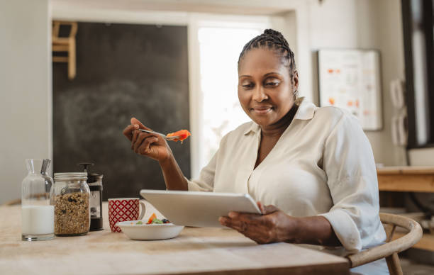 Smiling African American woman using a tablet and eating breakfast Smiling African American woman sitting at her kitchen table in the morning using a digital tablet while eating a healthy breakfast black people eating stock pictures, royalty-free photos & images