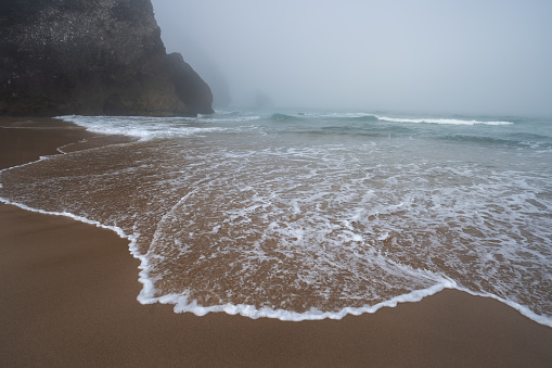 Praia da Adraga beach - White ocean waves and silhouette of costal rocks in morning fog. Sintra, Portugal.