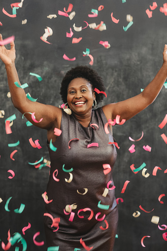 Portrait of a laughing mature African American woman wearing an evening gown tossing confetti up in the air in front of a chalkboard wall