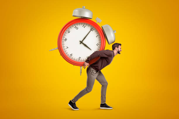 istock Side view of young handsome man carrying huge alarm clock on his back on yellow gradient background. 1179982226