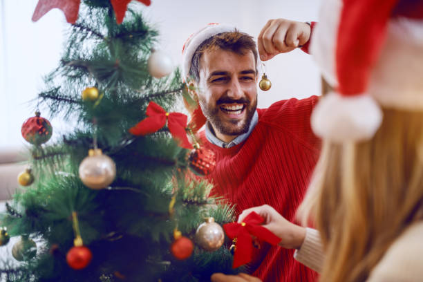 istock Handsome bearded smiling Caucasian man helping his girlfriend to decorate fir tree. Both having santa hats on heads. Living room interior. 1179987567