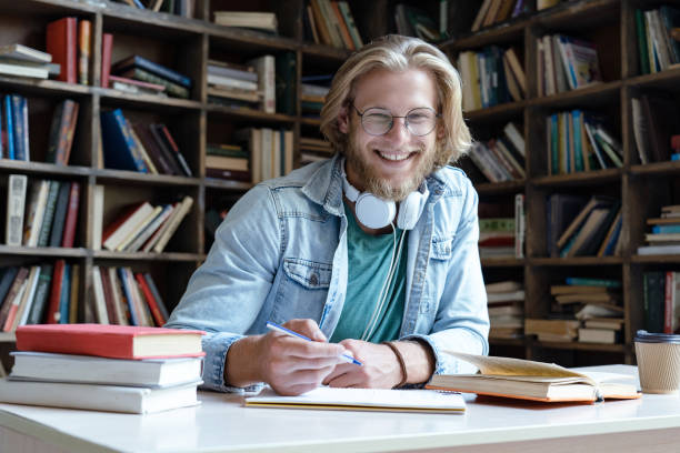 istock Happy guy university student in library learning with books headphones doing work assignment looking at camera sit at desk, smiling young man teacher wear glasses study write notes, portrait 1179989543