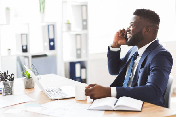 Black businessman talking on mobile phone, drinking coffee at office Smiling young black businessman talking on mobile phone, drinking coffee in modern white office, copy space businessman african descent on the phone business person stock pictures, royalty-free photos & images