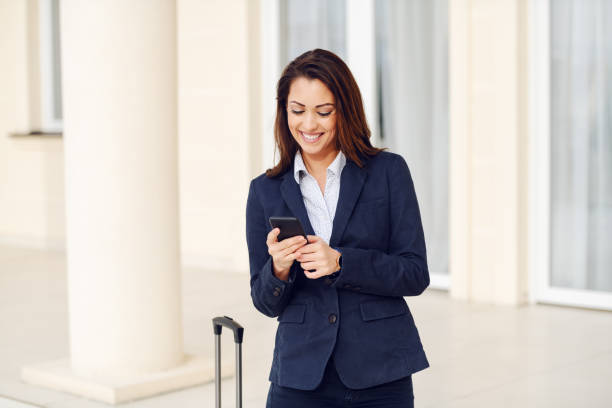istock Beautiful smiling caucasian brunette dressed smart casual using smart phone while standing outdoors. Next to woman is her luggage. Business trip concept. 1179991424