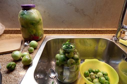 Preservation of green tomatoes at home. A large glass jar with green tomatoes stands in a stainless sink for dishes. On a granite countertop there is a closed can with tomatoes