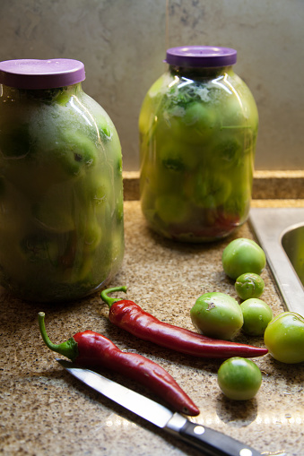Still life with red pepper. Two red hot peppers and green tomatoes lie on a granite countertop. In the background are two glass jars with green tomatoes.